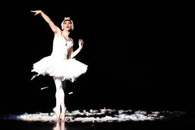 A male ballerina dressed in a white leotard and a feathered tutu poses en pointe on a black stage with a black background. Feathers are falling from the tutu and a pile of feathers surround the dancer on the floor.